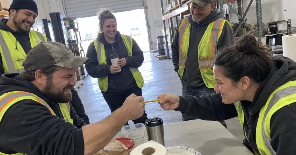 Coworkers in a warehouse break area watching two colleagues do a pencil grip ergonomics demo