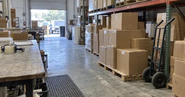 Warehouse aisle with heavy boxes stacked on a floor pallet and a high shelf, a hand truck and a packing table